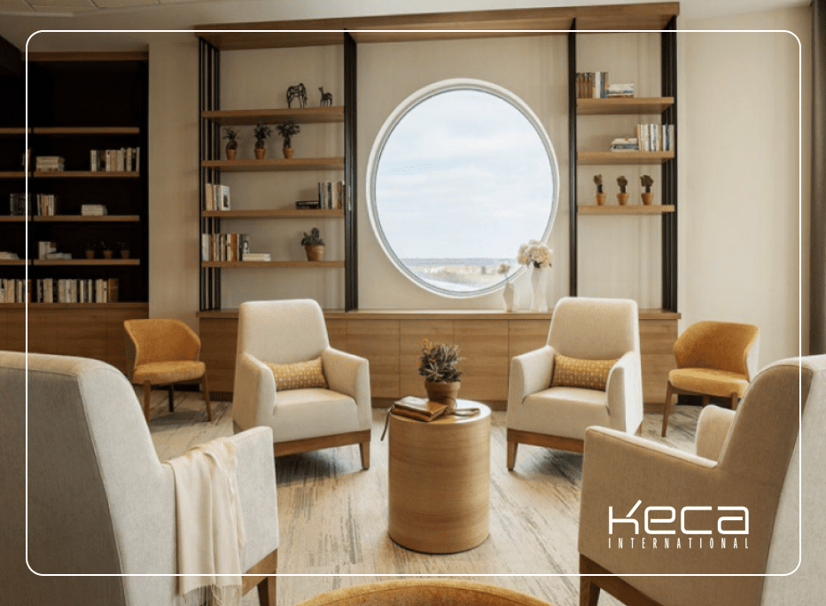 Warm senior living lounge featuring cream upholstered armchairs, a wooden side table, built-in bookshelves, and a large circular window with natural light, furnished by Keca International.