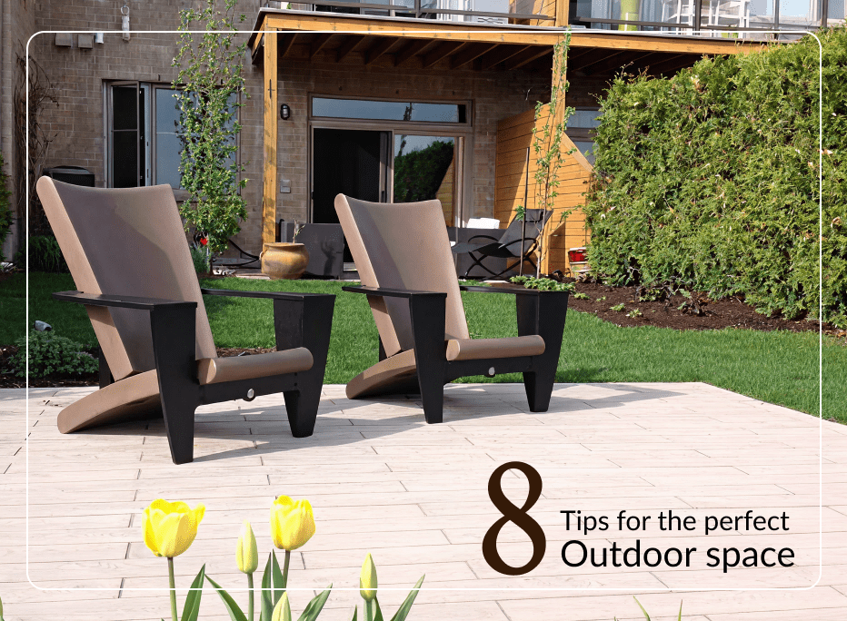 Contemporary outdoor lounge chairs with dark structural frames and taupe upholstered cushions arranged on a stone patio surrounded by lush landscaping and a modern building exterior.