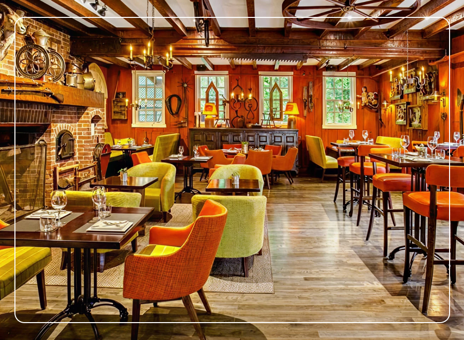 Warm rustic hotel restaurant dining room featuring orange and green upholstered commercial lounge chairs, wooden beam ceiling, brick fireplace, and bar stools creating an inviting hospitality atmosphere.