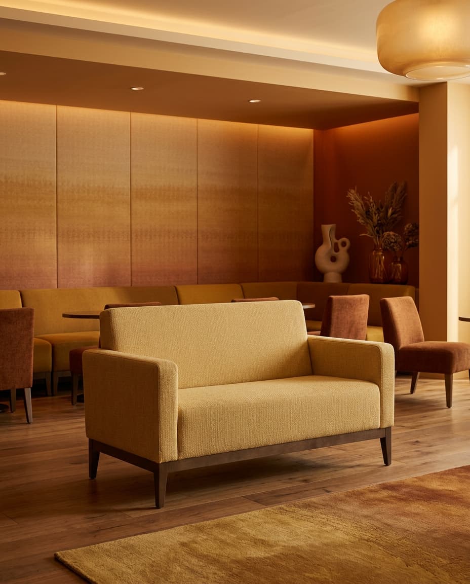 1850 two-seat sofa in textured ochre upholstery with a dark wood base and tapered legs, in a warmly lit hotel lounge with rust chairs and banquette seating behind.